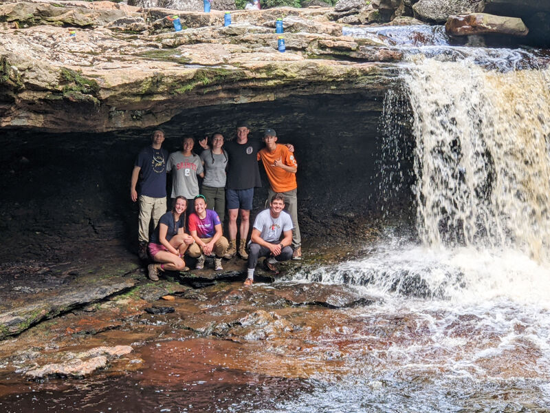 A group of nine people are posing for a picture in front of a waterfall. They are standing on rocks and under a rock overhang, with the waterfall cascading down to their right. The water is flowing rapidly, and the people are smiling and looking at the camera. The sky is not visible, but the lighting suggests it is daytime.